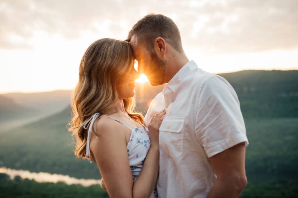 couple wearing white embraces at snooper's rock with sunrise behind them.